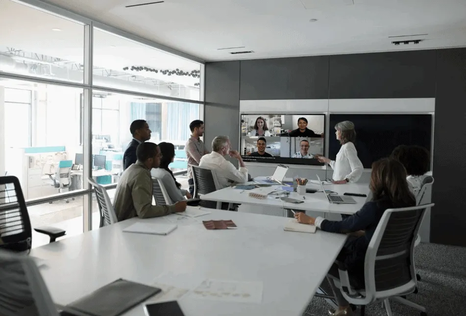 Seven people in a conference room gathered around a large table participating in a video conference with four other people visible on the large screen in the front of the room